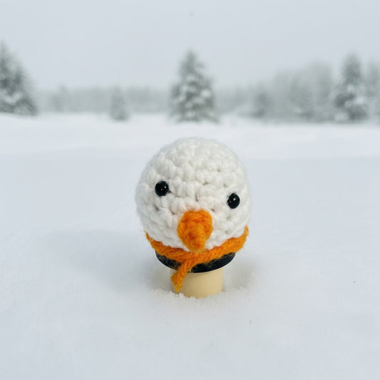 Crocheted snowman figure with an orange carrot nose on a bottle stopper in a snowy landscape