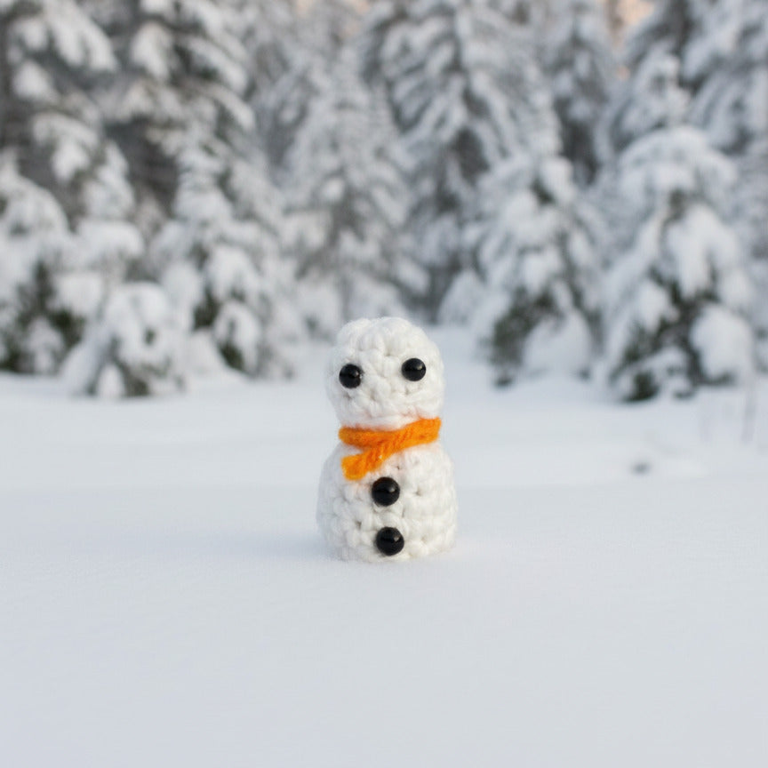 Small snowman figurine with an orange scarf in a snowy landscape with trees.