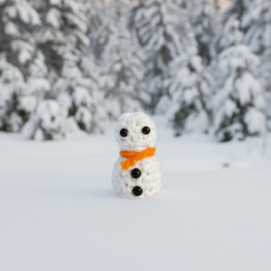 Small snowman figurine with an orange scarf in a snowy landscape with trees.